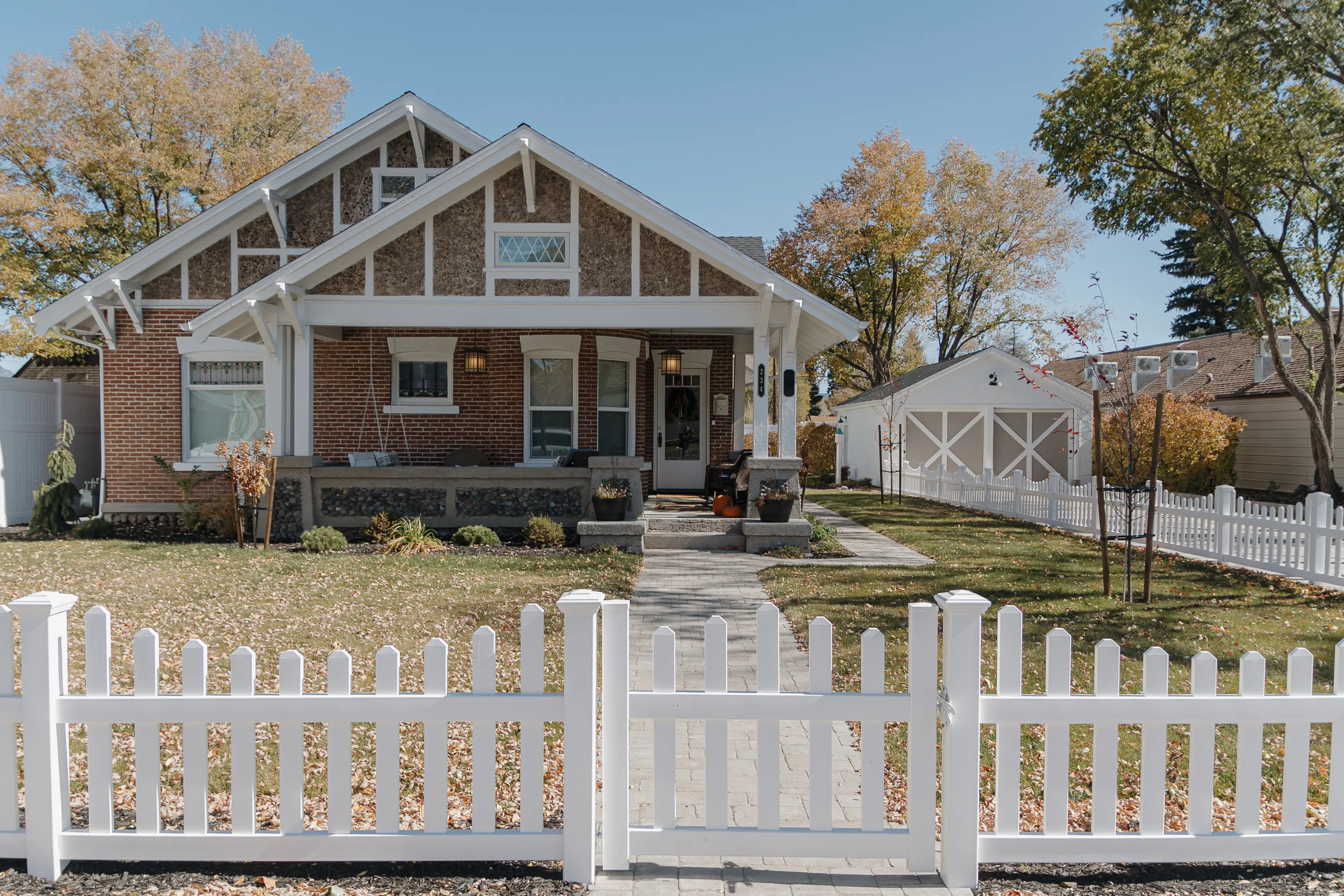 Front of a Craftsman-style brick bungalow with white trim, paved walkway, and white picket fence, leading to a detached garage.